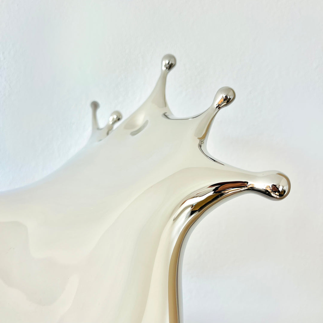 Polished stainless steel sculpture by Yoni Alter capturing the elegant moment of a liquid droplet mid-bounce. The reflective form twists fluidly to the left, with raised crown-like peaks suggesting impact and motion. A sculptural meditation on impermanence and form, grounded by a hidden counterweight for stability.