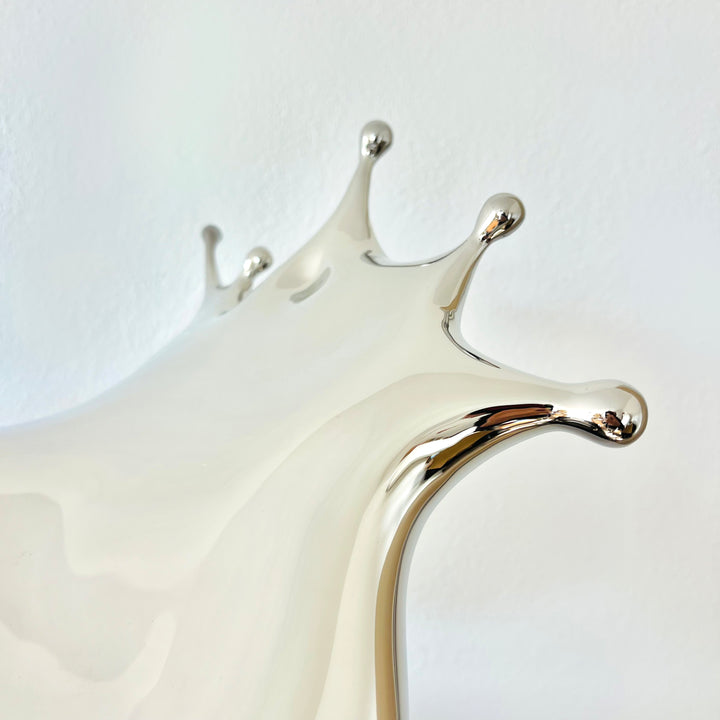 Polished stainless steel sculpture by Yoni Alter capturing the elegant moment of a liquid droplet mid-bounce. The reflective form twists fluidly to the left, with raised crown-like peaks suggesting impact and motion. A sculptural meditation on impermanence and form, grounded by a hidden counterweight for stability.