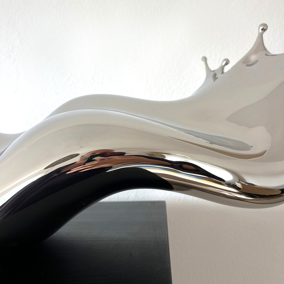 Polished stainless steel sculpture by Yoni Alter capturing the elegant moment of a liquid droplet mid-bounce. The reflective form twists fluidly to the left, with raised crown-like peaks suggesting impact and motion. A sculptural meditation on impermanence and form, grounded by a hidden counterweight for stability.