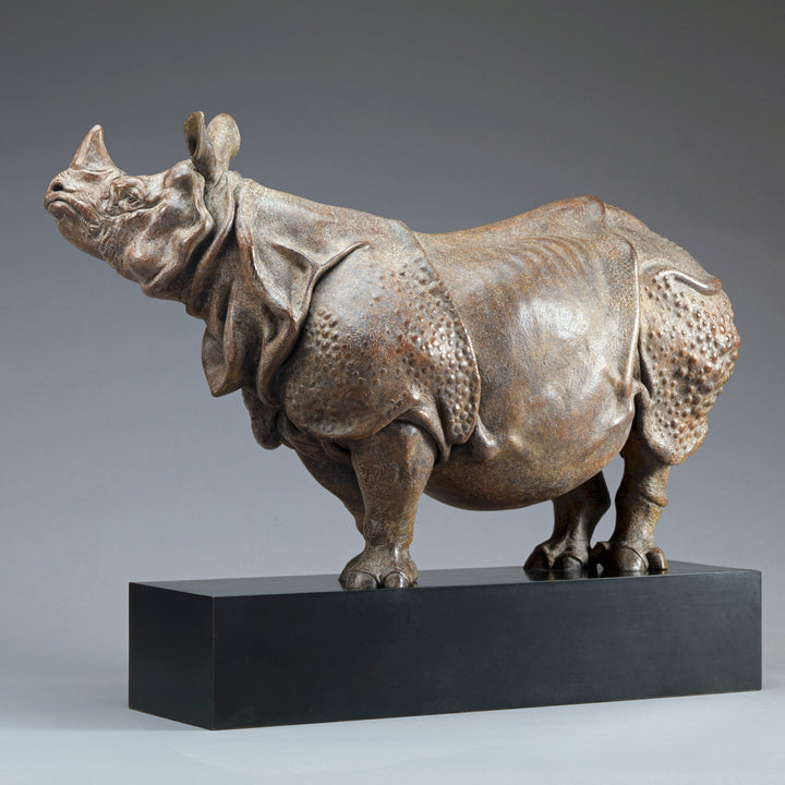 Large bronze Indian rhinoceros sculpture by Nick Bibby, showing armour-like skin folds, textured plates and powerful stance, finished in rich brown patina with subtle green tones on a polished stone plinth.