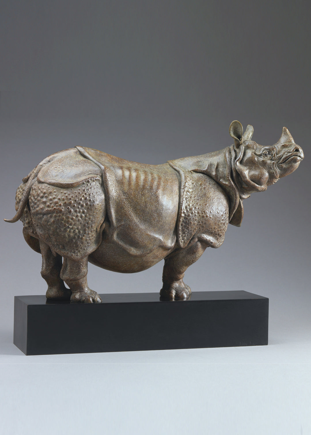 Large bronze Indian rhinoceros sculpture by Nick Bibby, showing armour-like skin folds, textured plates and powerful stance, finished in rich brown patina with subtle green tones on a polished stone plinth.