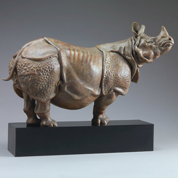 Large bronze Indian rhinoceros sculpture by Nick Bibby, showing armour-like skin folds, textured plates and powerful stance, finished in rich brown patina with subtle green tones on a polished stone plinth.