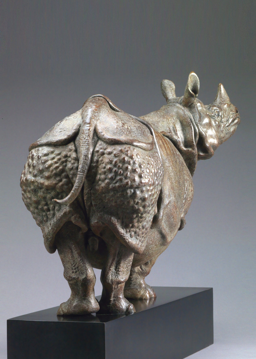 Large bronze Indian rhinoceros sculpture by Nick Bibby, showing armour-like skin folds, textured plates and powerful stance, finished in rich brown patina with subtle green tones on a polished stone plinth.