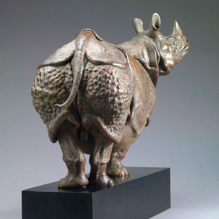 Large bronze Indian rhinoceros sculpture by Nick Bibby, showing armour-like skin folds, textured plates and powerful stance, finished in rich brown patina with subtle green tones on a polished stone plinth.