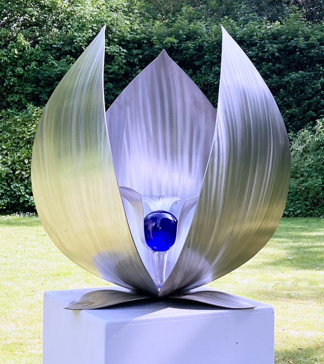Blue Seed by Ian Marlow — a stainless steel sculpture resembling a stylised flower or seed pod, featuring sweeping polished petals that cradle a vivid blue glass orb, set outdoors on a white plinth amidst lush greenery.