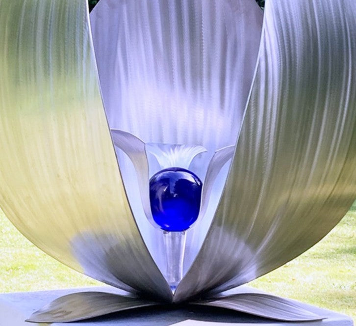 Blue Seed by Ian Marlow — a stainless steel sculpture resembling a stylised flower or seed pod, featuring sweeping polished petals that cradle a vivid blue glass orb, set outdoors on a white plinth amidst lush greenery.
