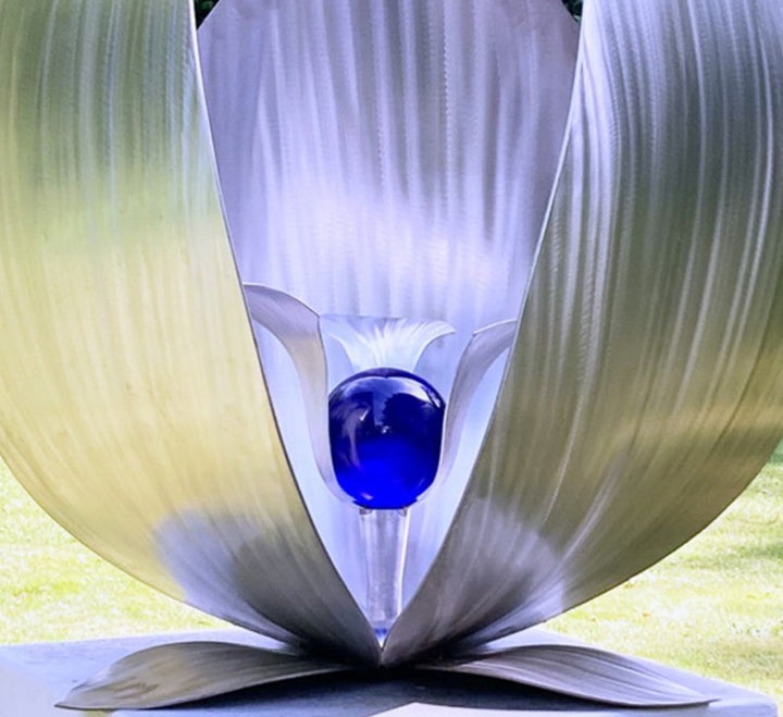 Blue Seed by Ian Marlow — a stainless steel sculpture resembling a stylised flower or seed pod, featuring sweeping polished petals that cradle a vivid blue glass orb, set outdoors on a white plinth amidst lush greenery.