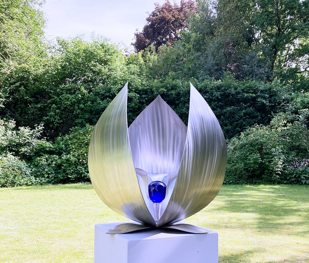 Blue Seed by Ian Marlow — a stainless steel sculpture resembling a stylised flower or seed pod, featuring sweeping polished petals that cradle a vivid blue glass orb, set outdoors on a white plinth amidst lush greenery.