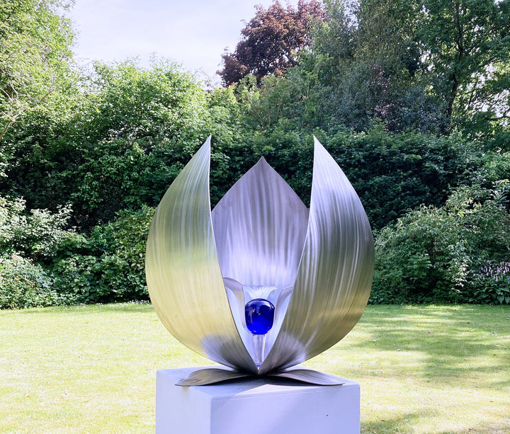 Blue Seed by Ian Marlow — a stainless steel sculpture resembling a stylised flower or seed pod, featuring sweeping polished petals that cradle a vivid blue glass orb, set outdoors on a white plinth amidst lush greenery.