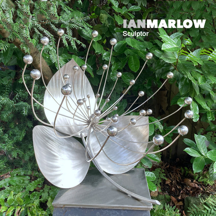 A stainless steel sculpture titled Flower by Ian Marlow, featuring sweeping metallic petals and radiating stems tipped with polished spheres, presented outdoors on a square plinth among rich garden foliage.