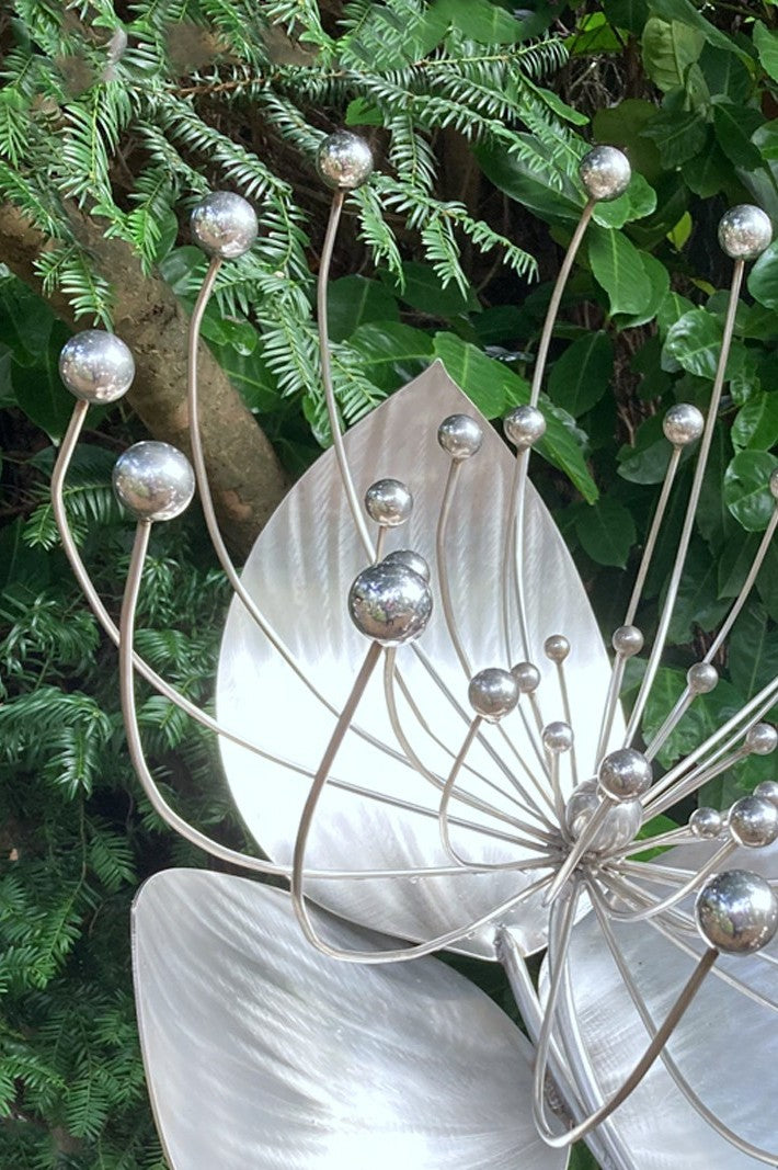 A stainless steel sculpture titled Flower by Ian Marlow, featuring sweeping metallic petals and radiating stems tipped with polished spheres, presented outdoors on a square plinth among rich garden foliage.