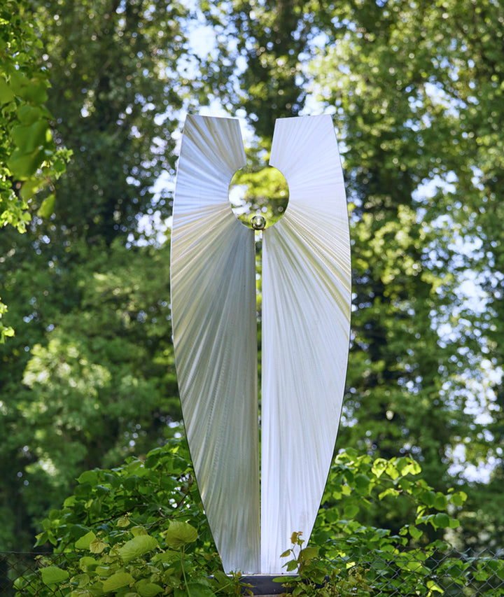Light-Bringer I by Ian Marlow — a vertically split stainless steel sculpture with dynamic radial surface textures and a central circular void holding a polished sphere, set outdoors among vibrant greenery.