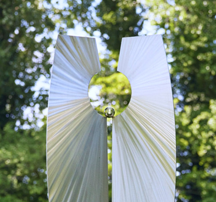 Light-Bringer I by Ian Marlow — a vertically split stainless steel sculpture with dynamic radial surface textures and a central circular void holding a polished sphere, set outdoors among vibrant greenery.