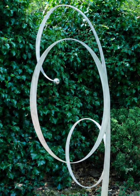 Ian Marlow MRSS stainless steel sculpture Orbiting Upwards, featuring a tall contemporary abstract form with sweeping interconnected circular loops rising upwards, displayed outdoors within a landscaped garden setting.