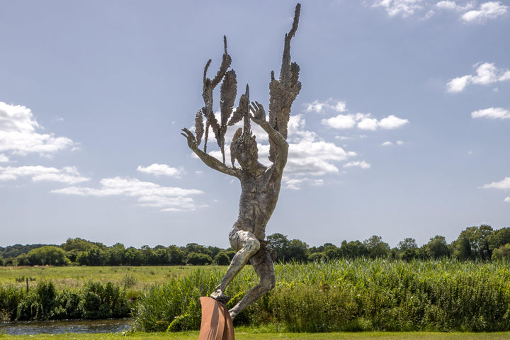 Monumental outdoor bronze sculpture by Nicola Godden — a dramatic, winged male figure with raised arms, climbing and balancing on a sweeping rusted steel crescent. The figure’s body has a textured verdigris green and earthy bronze patina with highlights of gold and brown, contrasting with the deep weathered red-orange steel base. Photographed in an open grassy field with blue sky.