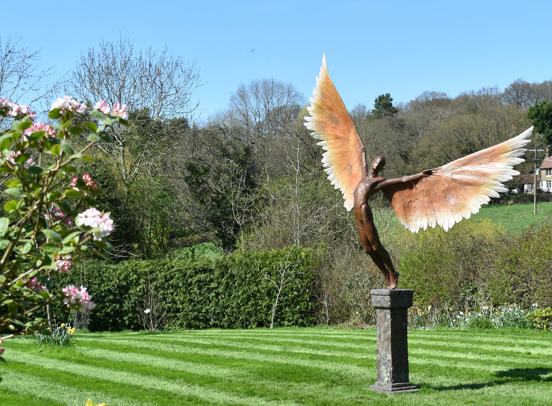 Icarus I — monumental bronze sculpture by Nicola Godden, depicting a stylised male figure with immense outstretched wings, leaning forward as if preparing for flight. Rich bronze patina with warm coppery browns fading into lighter golden tips across the feathered wings. Originally featured at the London 2012 Olympic Village and sold at Sotheby’s. Set outdoors against a clear blue sky and classical architectural backdrop.