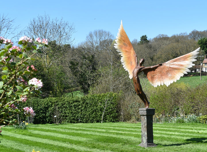 Icarus I — monumental bronze sculpture by Nicola Godden, depicting a stylised male figure with immense outstretched wings, leaning forward as if preparing for flight. Rich bronze patina with warm coppery browns fading into lighter golden tips across the feathered wings. Originally featured at the London 2012 Olympic Village and sold at Sotheby’s. Set outdoors against a clear blue sky and classical architectural backdrop.