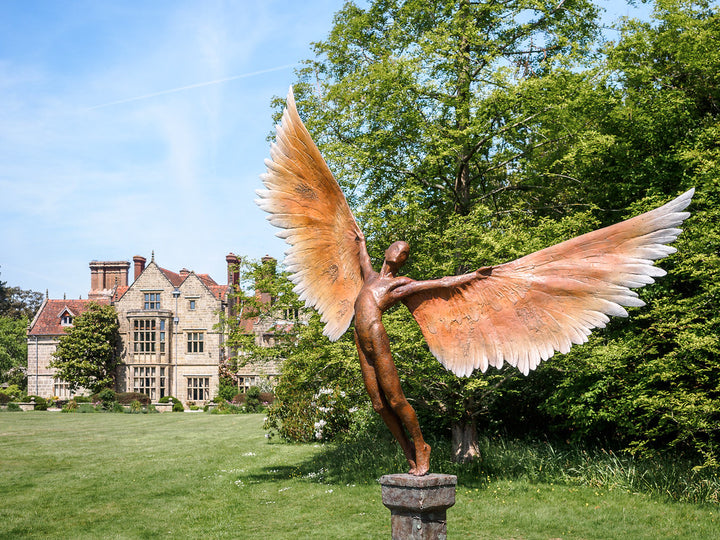Icarus I — monumental bronze sculpture by Nicola Godden, depicting a stylised male figure with immense outstretched wings, leaning forward as if preparing for flight. Rich bronze patina with warm coppery browns fading into lighter golden tips across the feathered wings. Originally featured at the London 2012 Olympic Village and sold at Sotheby’s. Set outdoors against a clear blue sky and classical architectural backdrop.