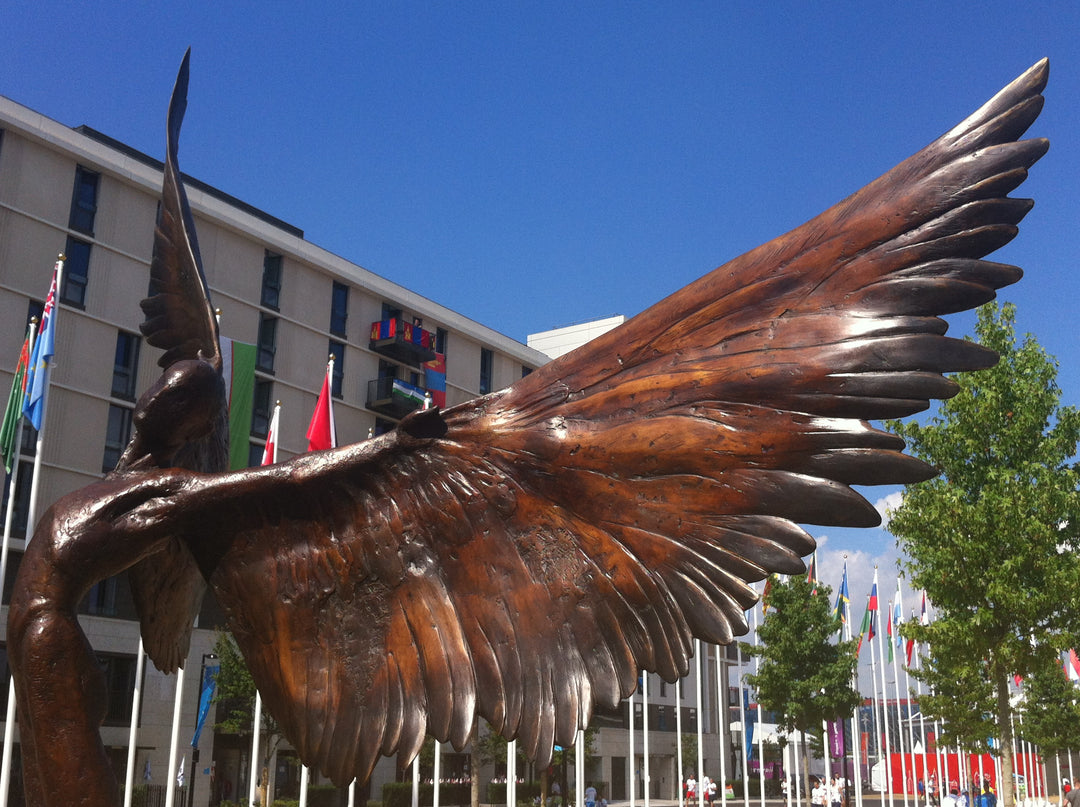 Icarus I — monumental bronze sculpture by Nicola Godden, depicting a stylised male figure with immense outstretched wings, leaning forward as if preparing for flight. Rich bronze patina with warm coppery browns fading into lighter golden tips across the feathered wings. Originally featured at the London 2012 Olympic Village and sold at Sotheby’s. Set outdoors against a clear blue sky and classical architectural backdrop.