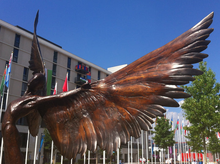 Icarus I — monumental bronze sculpture by Nicola Godden, depicting a stylised male figure with immense outstretched wings, leaning forward as if preparing for flight. Rich bronze patina with warm coppery browns fading into lighter golden tips across the feathered wings. Originally featured at the London 2012 Olympic Village and sold at Sotheby’s. Set outdoors against a clear blue sky and classical architectural backdrop.