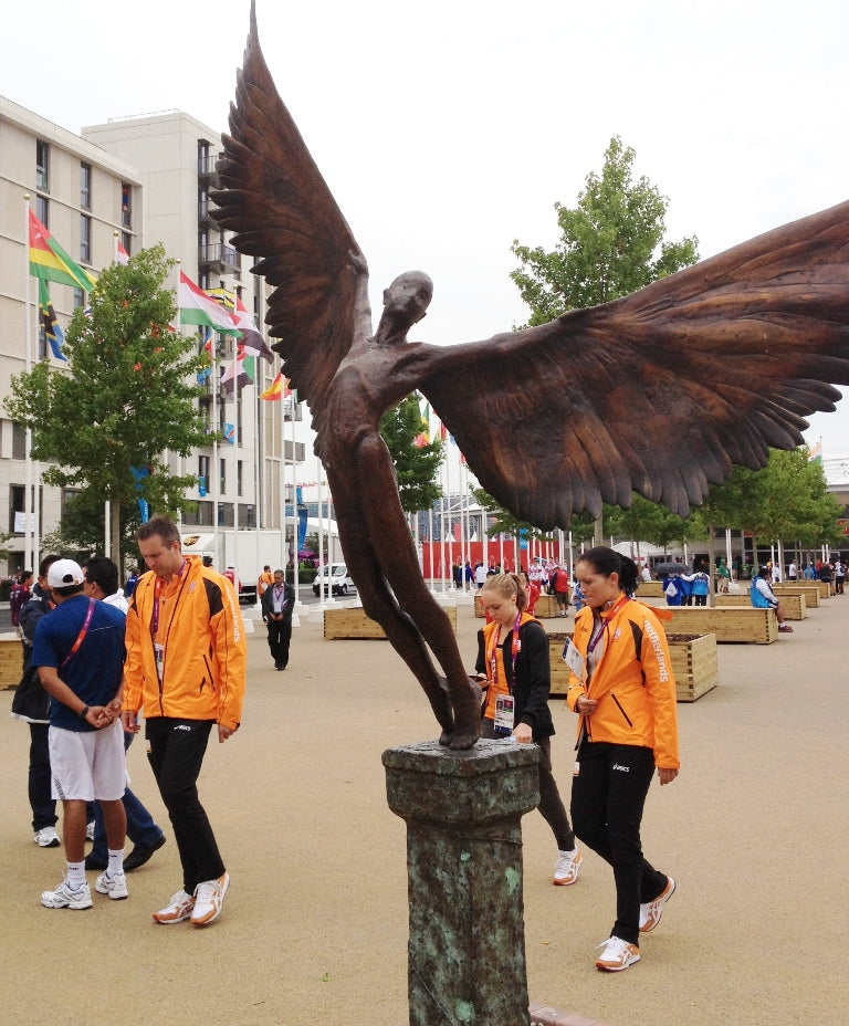 Icarus I — monumental bronze sculpture by Nicola Godden, depicting a stylised male figure with immense outstretched wings, leaning forward as if preparing for flight. Rich bronze patina with warm coppery browns fading into lighter golden tips across the feathered wings. Originally featured at the London 2012 Olympic Village and sold at Sotheby’s. Set outdoors against a clear blue sky and classical architectural backdrop.