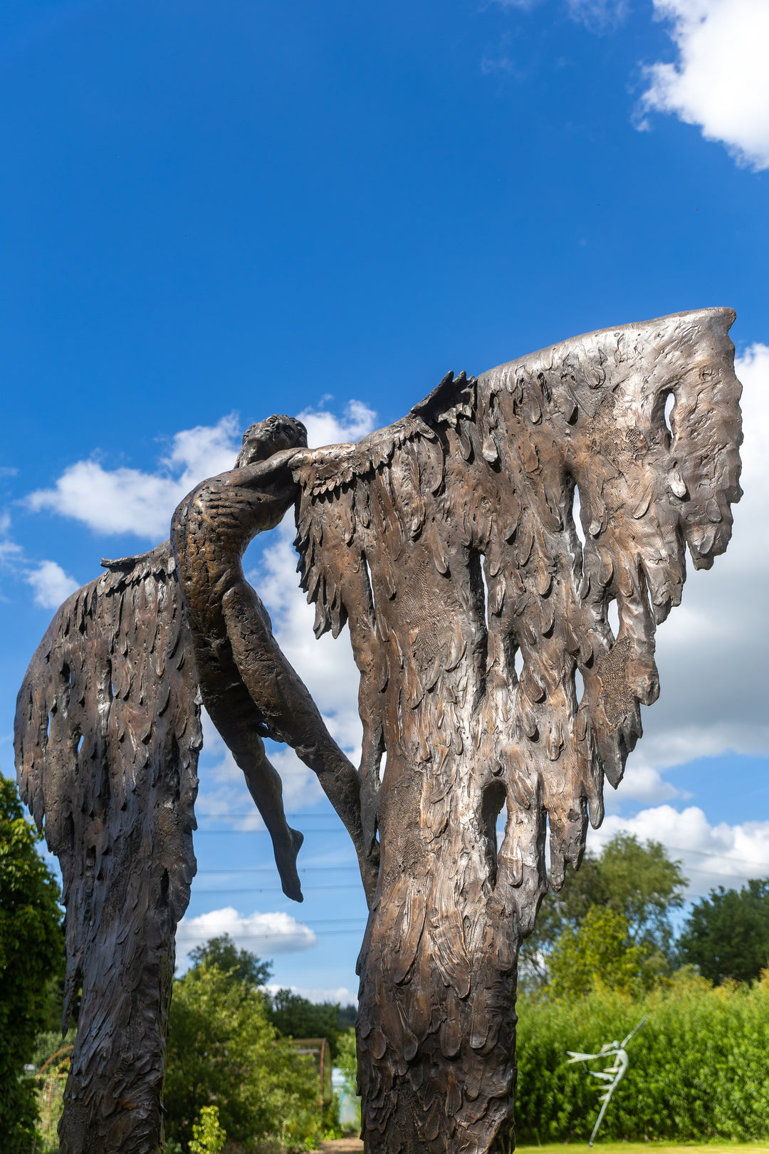Monumental bronze sculpture of Icarus mid-flight with expansive feathered wings, captured in an expressive upward pose against a bright sky and lush garden backdrop.