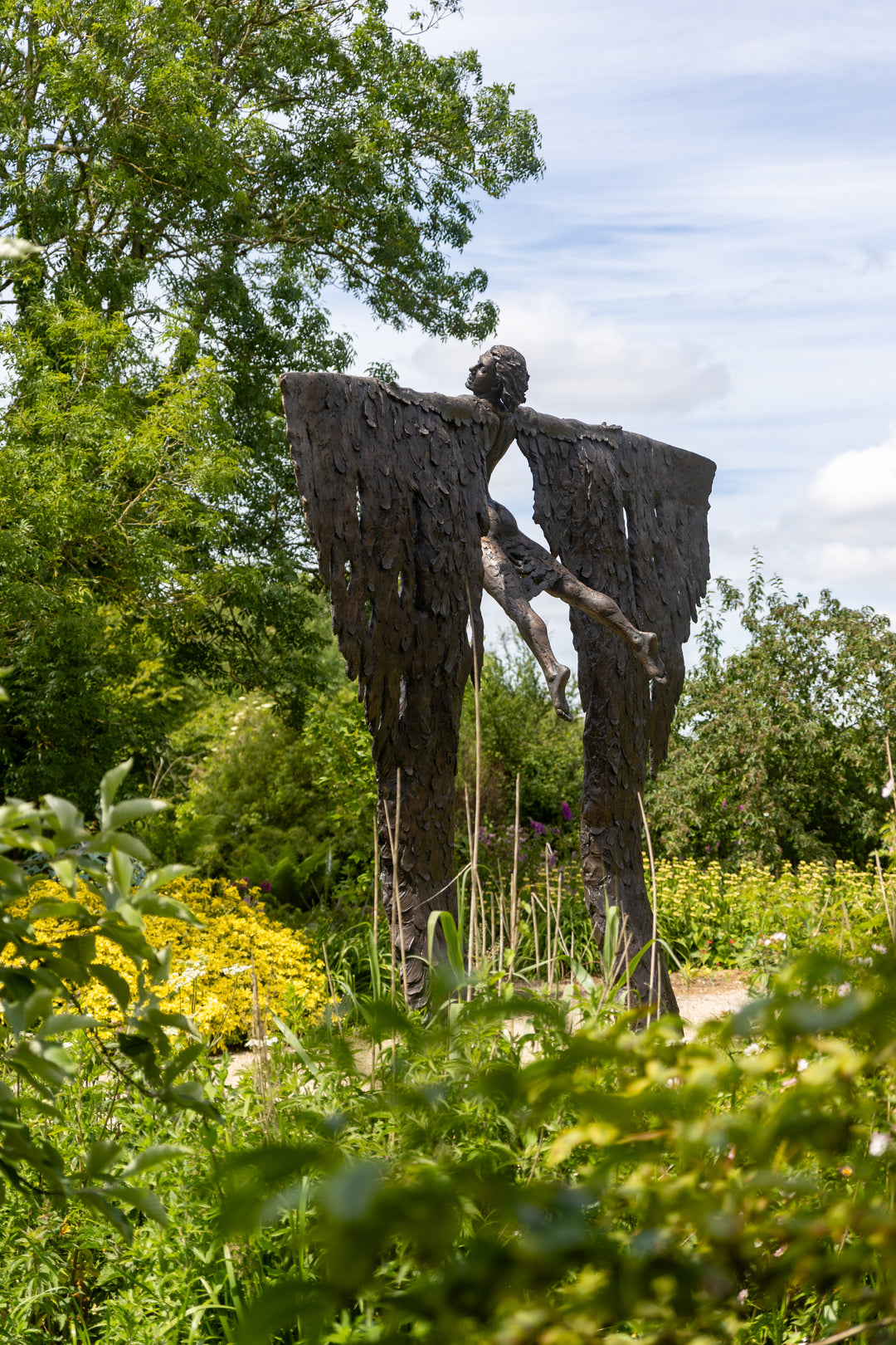 Monumental bronze sculpture of Icarus mid-flight with expansive feathered wings, captured in an expressive upward pose against a bright sky and lush garden backdrop.