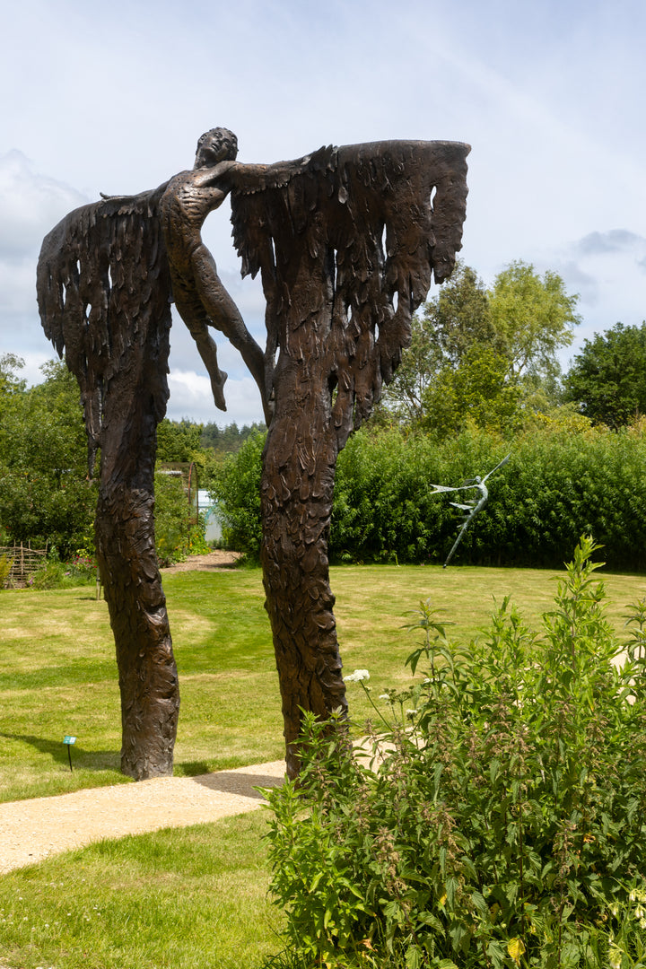 Monumental bronze sculpture of Icarus mid-flight with expansive feathered wings, captured in an expressive upward pose against a bright sky and lush garden backdrop.