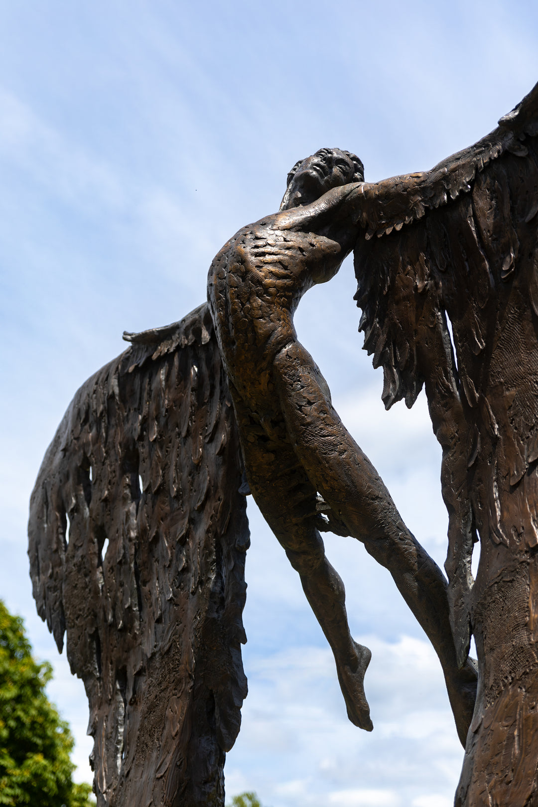 Monumental bronze sculpture of Icarus mid-flight with expansive feathered wings, captured in an expressive upward pose against a bright sky and lush garden backdrop.
