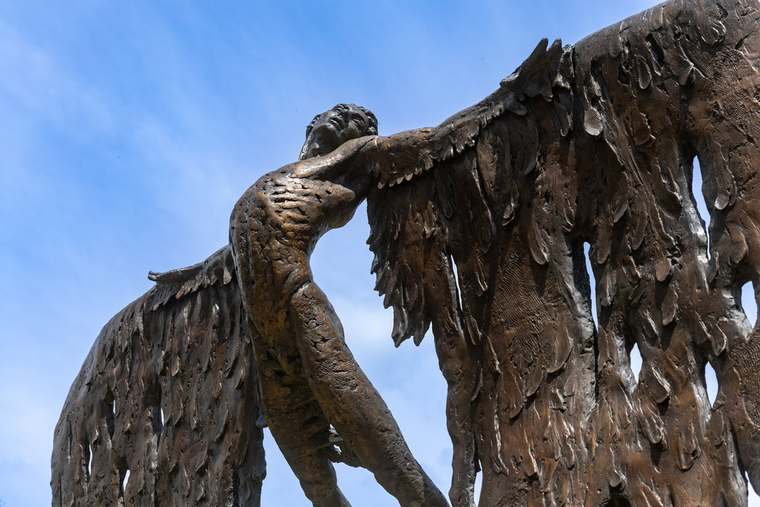 Monumental bronze sculpture of Icarus mid-flight with expansive feathered wings, captured in an expressive upward pose against a bright sky and lush garden backdrop.