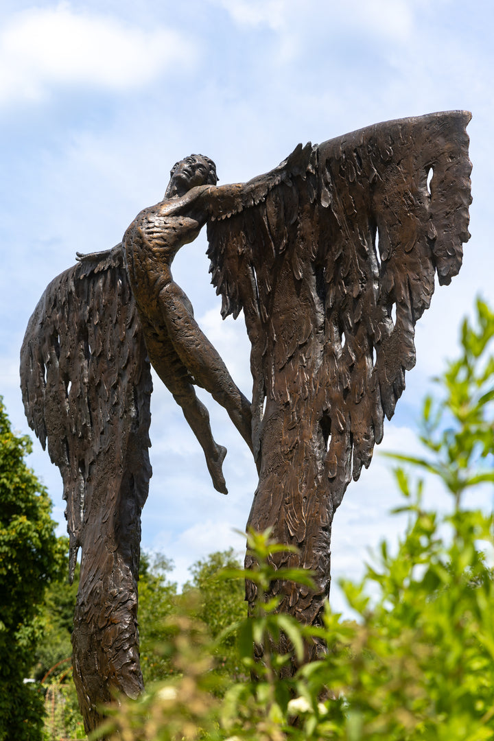 Monumental bronze sculpture of Icarus mid-flight with expansive feathered wings, captured in an expressive upward pose against a bright sky and lush garden backdrop.