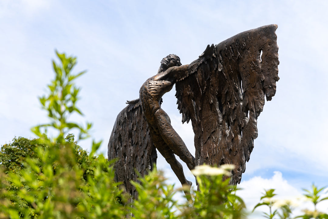 Monumental bronze sculpture of Icarus mid-flight with expansive feathered wings, captured in an expressive upward pose against a bright sky and lush garden backdrop.