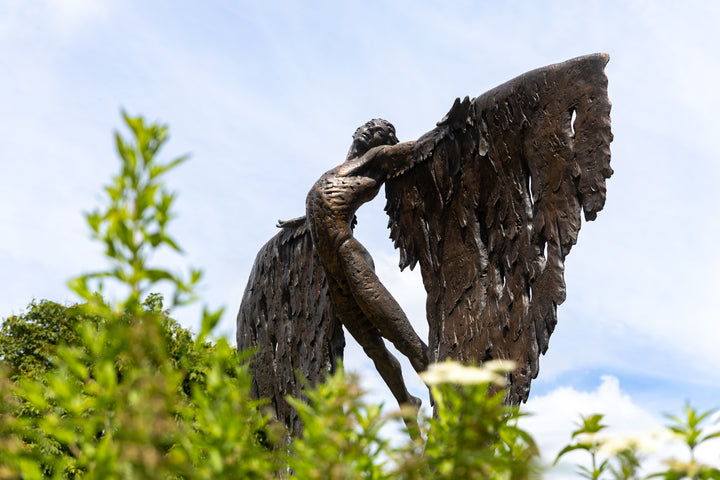 Monumental bronze sculpture of Icarus mid-flight with expansive feathered wings, captured in an expressive upward pose against a bright sky and lush garden backdrop.