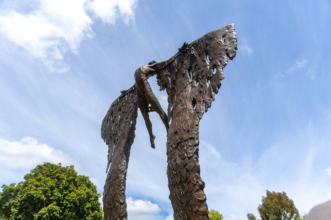 Monumental bronze sculpture of Icarus mid-flight with expansive feathered wings, captured in an expressive upward pose against a bright sky and lush garden backdrop.