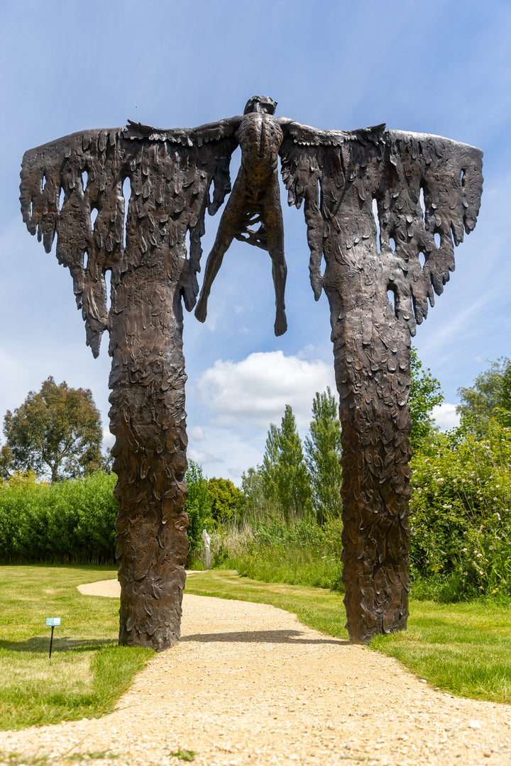 Monumental bronze sculpture of Icarus mid-flight with expansive feathered wings, captured in an expressive upward pose against a bright sky and lush garden backdrop.