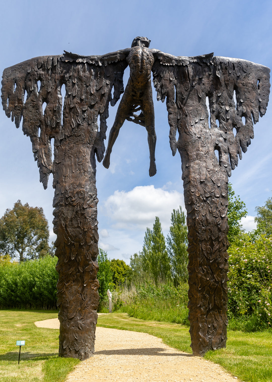 Monumental bronze sculpture of Icarus mid-flight with expansive feathered wings, captured in an expressive upward pose against a bright sky and lush garden backdrop.