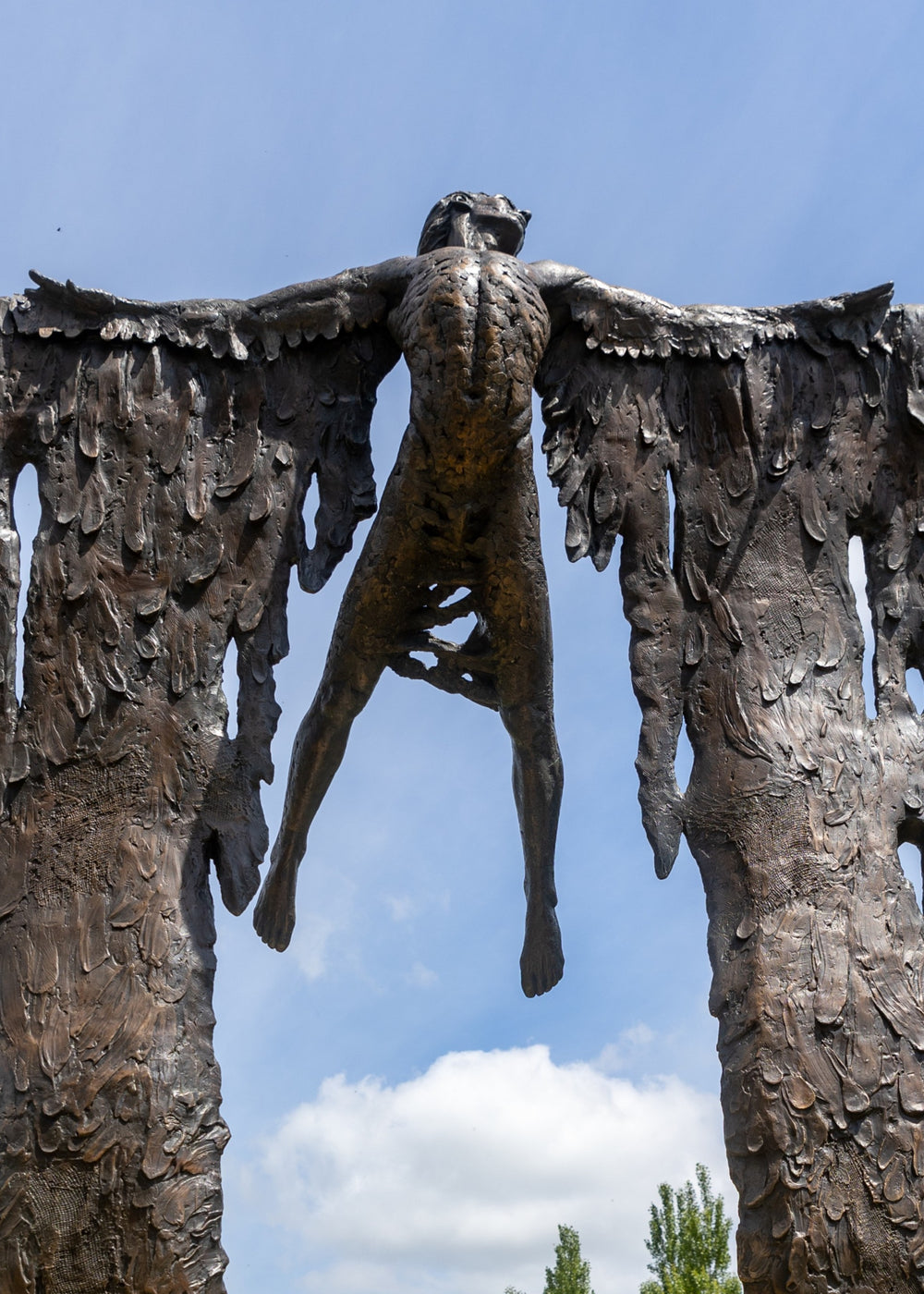 Monumental bronze sculpture of Icarus mid-flight with expansive feathered wings, captured in an expressive upward pose against a bright sky and lush garden backdrop.