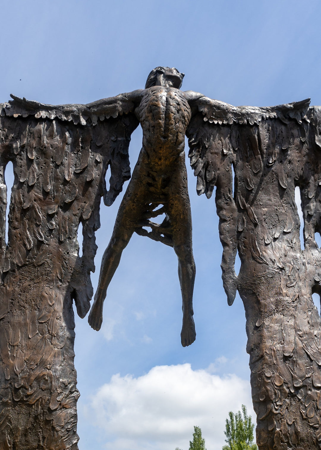 Monumental bronze sculpture of Icarus mid-flight with expansive feathered wings, captured in an expressive upward pose against a bright sky and lush garden backdrop.