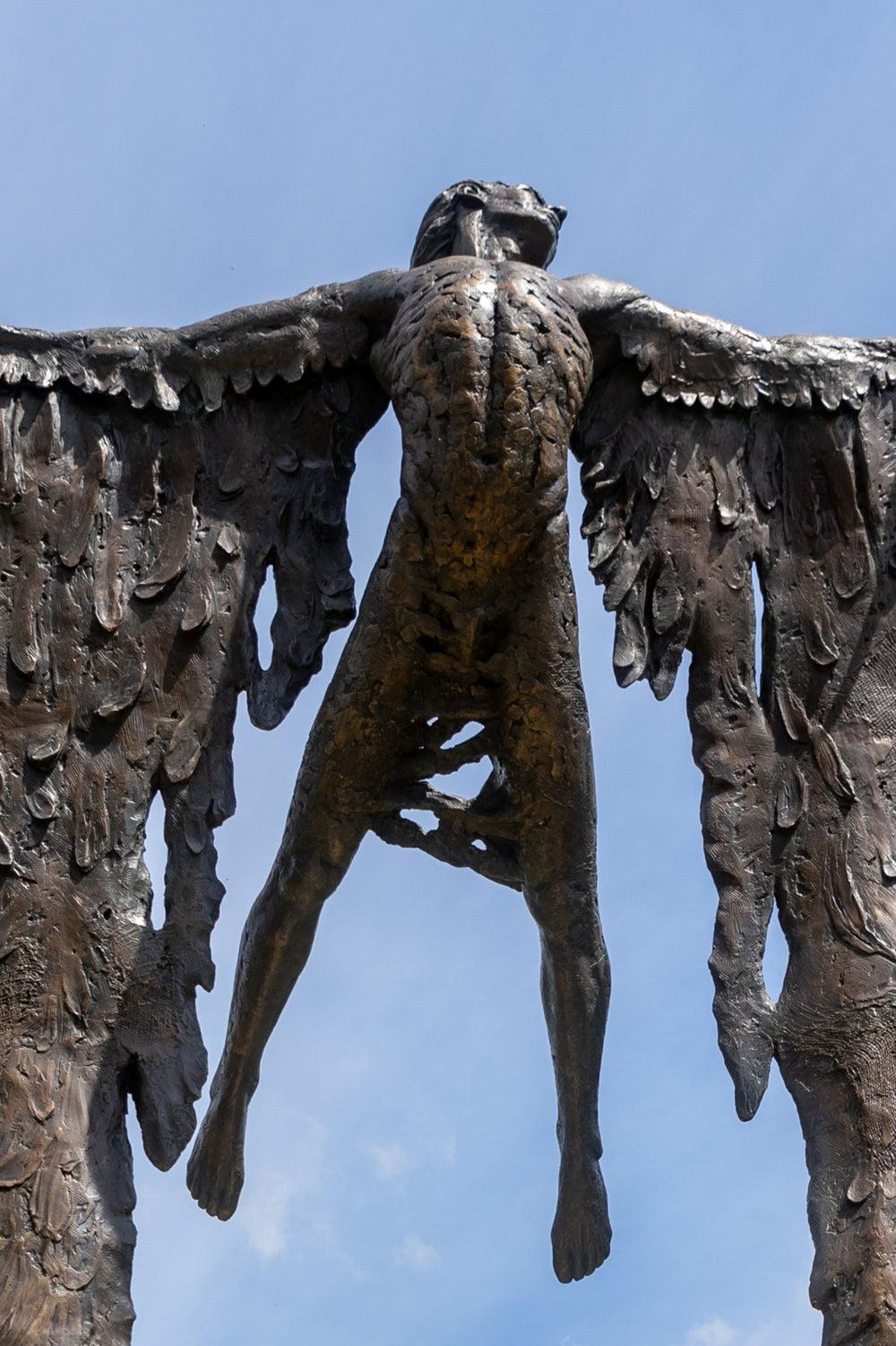 Monumental bronze sculpture of Icarus mid-flight with expansive feathered wings, captured in an expressive upward pose against a bright sky and lush garden backdrop.
