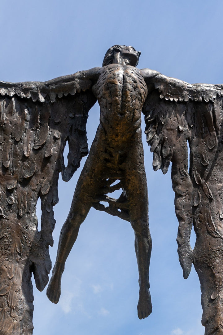 Monumental bronze sculpture of Icarus mid-flight with expansive feathered wings, captured in an expressive upward pose against a bright sky and lush garden backdrop.