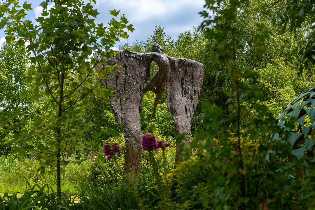 Monumental bronze sculpture of Icarus mid-flight with expansive feathered wings, captured in an expressive upward pose against a bright sky and lush garden backdrop.
