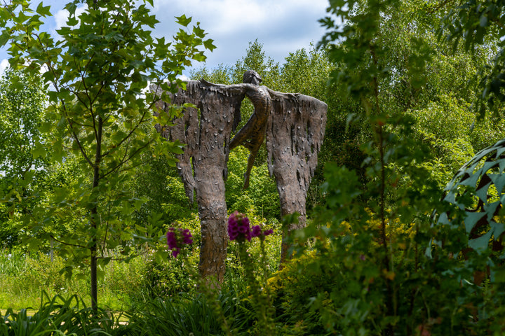 Monumental bronze sculpture of Icarus mid-flight with expansive feathered wings, captured in an expressive upward pose against a bright sky and lush garden backdrop.