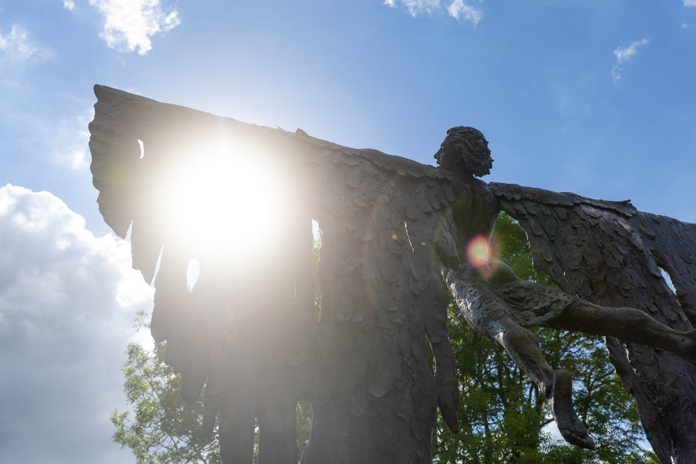 Monumental bronze sculpture of Icarus mid-flight with expansive feathered wings, captured in an expressive upward pose against a bright sky and lush garden backdrop.