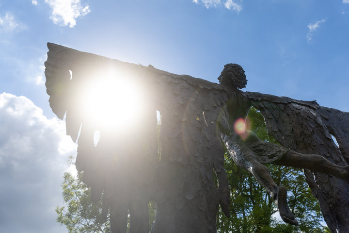 Monumental bronze sculpture of Icarus mid-flight with expansive feathered wings, captured in an expressive upward pose against a bright sky and lush garden backdrop.