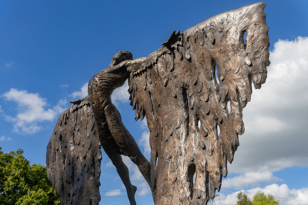 Monumental bronze sculpture of Icarus mid-flight with expansive feathered wings, captured in an expressive upward pose against a bright sky and lush garden backdrop.