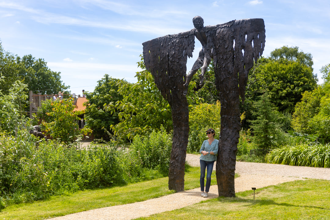 Monumental bronze sculpture of Icarus mid-flight with expansive feathered wings, captured in an expressive upward pose against a bright sky and lush garden backdrop.