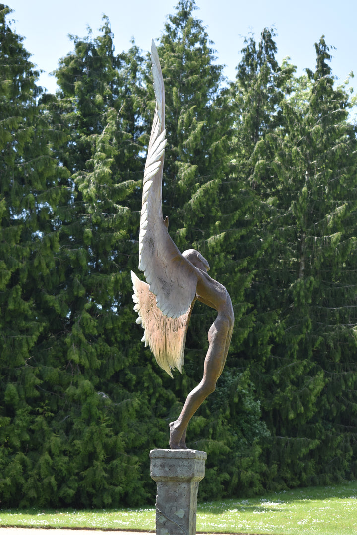 Icarus I — monumental bronze sculpture by Nicola Godden, depicting a stylised male figure with immense outstretched wings, leaning forward as if preparing for flight. Rich bronze patina with warm coppery browns fading into lighter golden tips across the feathered wings. Originally featured at the London 2012 Olympic Village and sold at Sotheby’s. Set outdoors against a clear blue sky and classical architectural backdrop.