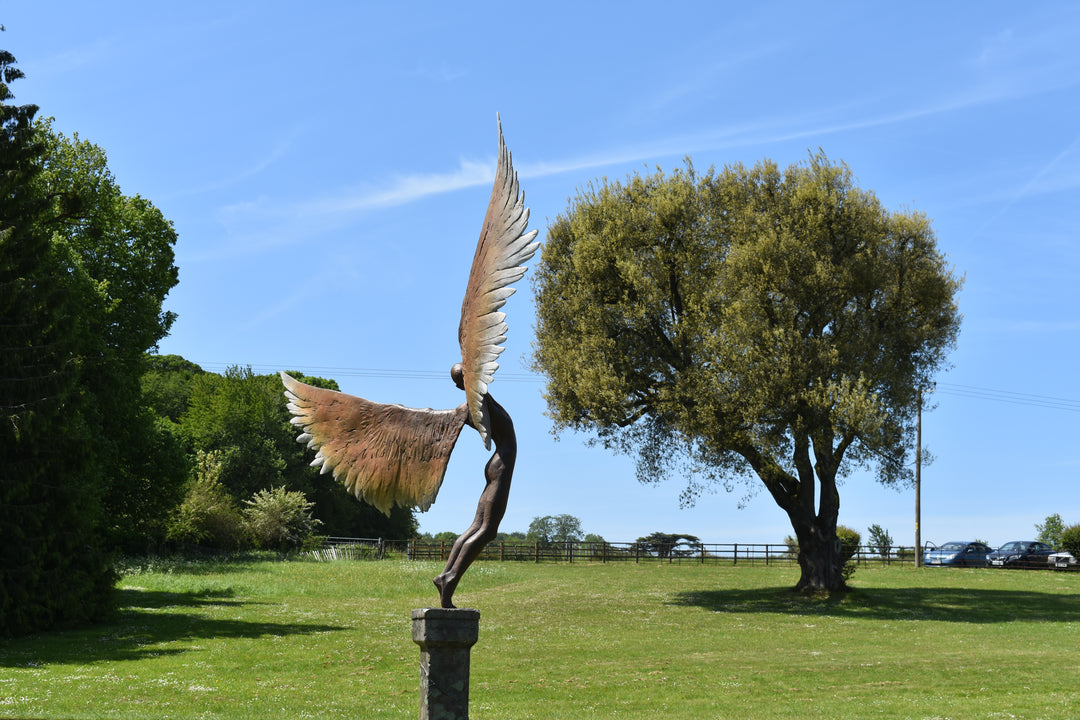 Icarus I — monumental bronze sculpture by Nicola Godden, depicting a stylised male figure with immense outstretched wings, leaning forward as if preparing for flight. Rich bronze patina with warm coppery browns fading into lighter golden tips across the feathered wings. Originally featured at the London 2012 Olympic Village and sold at Sotheby’s. Set outdoors against a clear blue sky and classical architectural backdrop.