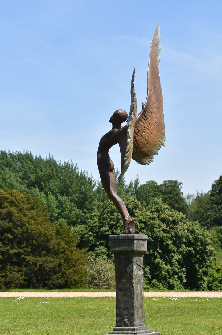 Icarus I — monumental bronze sculpture by Nicola Godden, depicting a stylised male figure with immense outstretched wings, leaning forward as if preparing for flight. Rich bronze patina with warm coppery browns fading into lighter golden tips across the feathered wings. Originally featured at the London 2012 Olympic Village and sold at Sotheby’s. Set outdoors against a clear blue sky and classical architectural backdrop.