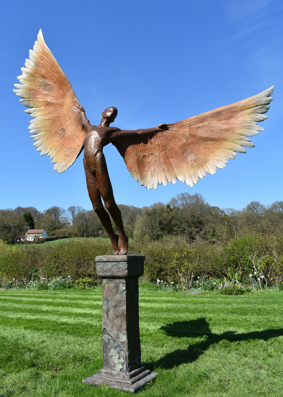 Icarus I — monumental bronze sculpture by Nicola Godden, depicting a stylised male figure with immense outstretched wings, leaning forward as if preparing for flight. Rich bronze patina with warm coppery browns fading into lighter golden tips across the feathered wings. Originally featured at the London 2012 Olympic Village and sold at Sotheby’s. Set outdoors against a clear blue sky and classical architectural backdrop.